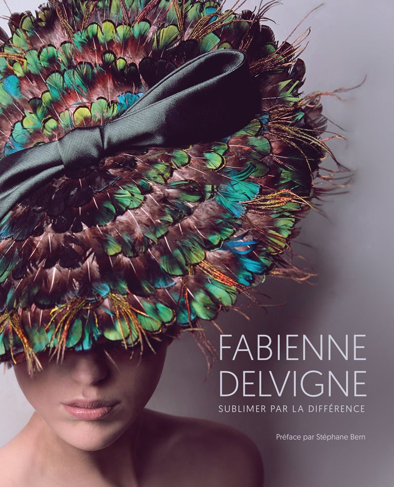 Fashion model in brown, blue and green feathered hat with black silk bow to centre, FABIENNE DELVIGNE in white font to lower right.