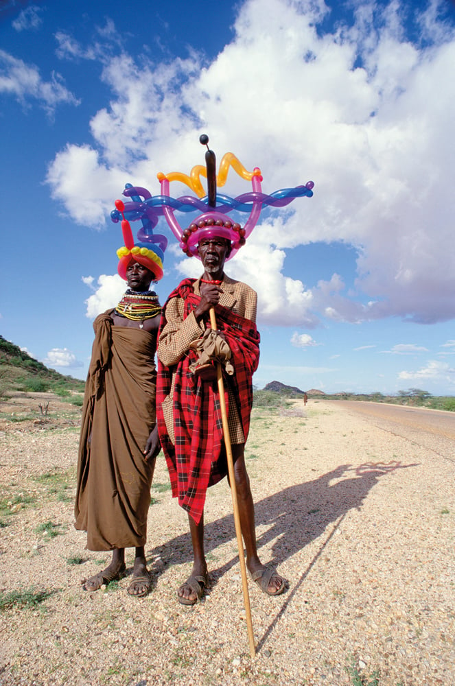 2 African men in traditional dress, one leaning on stick, wearing inflated balloon crowns on their heads.