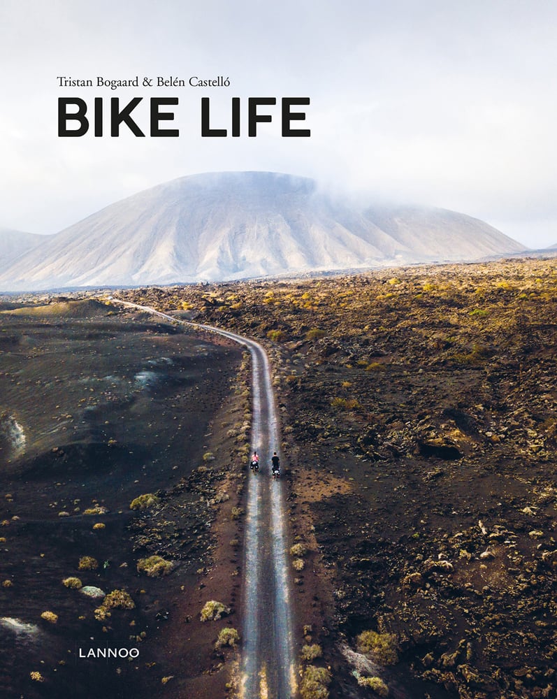 Aerial view of 2 people on bicycles riding on road towards mountainous landscape, on cover of 'Bike Life, Travel, Different', by Lannoo Publishers.