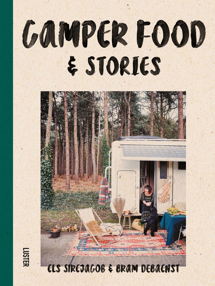 Woman sitting in doorway of campervan with black dog, in forest area, on cover of 'Camper Food & Stories', by Luster Publishing.