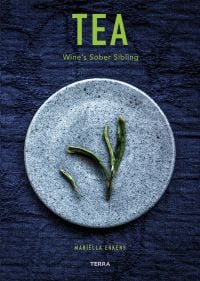 Green tea leaves on pale blue plate, deep blue tablecloth beneath, on cover of 'Tea', Wine's Sober Sibling', by Lannoo Publishers.