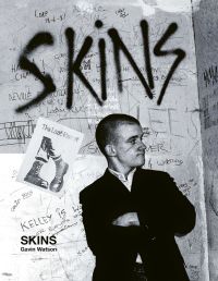 Young white male skinhead wearing black jacket, arms folded, standing in front of wall graffitied with 'SKINS', in black spray paint, by ACC Art Books.