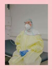 Pink book cover of Ingmar Björn Nolting, About the Days Ahead, featuring person in yellow medical gown, blue gloves face mask and goggles, sitting on hospital bed. Published by Verlag Kettler.