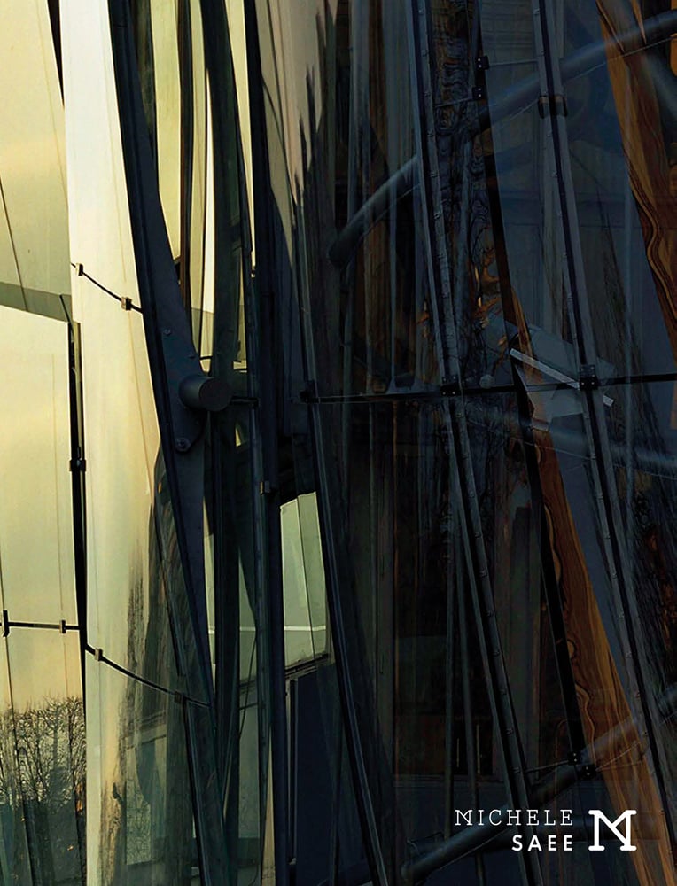 Close up of building windows of metal structure, reflections of trees in glass, Michele Saee in white font to bottom right.