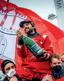 Boris Hermann smiling while on boat in red waterproofs holding back sail with one hand, on cover of 'Nonstop, Süchtig nach Segeln / Driven by the sea', by Delius Klasing Verlag GmbH.