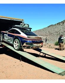 Book cover of Rough Roads to 911 Dakar, featuring a black Porsche 911 racing down a sand dune in the desert. Published by Delius.