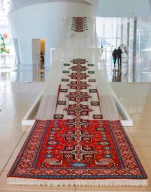 Book cover of Faig Ahmed, with a large, red textile rug suspended on white exhibition wall, with the black bottom half in shape of spilt liquid. Published by Kerber.