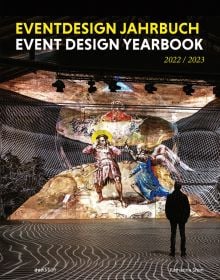 Book cover of Event Design Yearbook 2022 / 2023, with an exhibition space, and a man looking at figure with wings fighting figure with sword. Published by Avedition Gmbh.